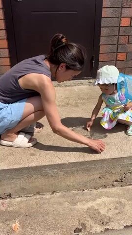 A woman and a child are outside. The woman is wearing a grey tank top and blue shorts. She is kneeling down and pointing at something on the ground. The child is wearing a white hat, a colorful dress, and sandals. The child is also kneeling and appears to be holding a blue net. They are on a concrete surface in front of a dark brown door and a brick wall. The sun is shining, casting shadows on the ground.