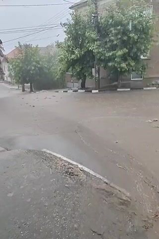 A video taken from a low angle shows a flooded street during a rainstorm. The street is covered in a thin layer of water, and the curb in the foreground is visible. Buildings and trees line the street in the background. The camera is stationary for the entire duration of the clip.