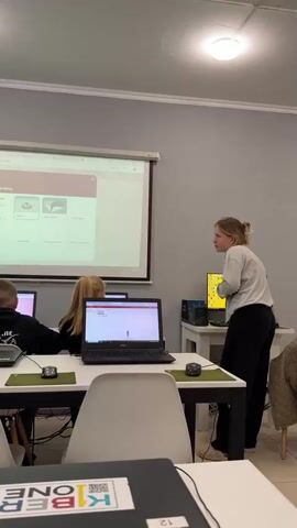 The video shows a classroom setting where a teacher is interacting with students who are seated at computers. The teacher, a young woman with blonde hair, is standing and gesturing towards a projector screen. The screen displays a webpage with a title that appears to be in Russian and shows various icons or options. Several students, including children and possibly teenagers, are facing their laptops, which are open and on. Some of the students are looking at their screens, while others are looking towards the teacher or the projector. The classroom environment appears to be well-lit with fluorescent ceiling lights and has grey walls. There are white tables where the students are working, and white chairs are visible. A green curtain is on the left side of the room. The overall atmosphere suggests a lesson or a workshop involving computers and digital content.