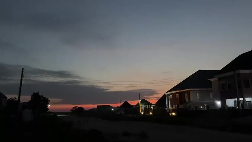 The video presents a static, low-angle shot of a residential street or community at dusk, with the sky displaying a gradient of red-orange near the horizon blending into dark blue and gray clouds overhead. Several multi-story houses with illuminated lights are visible on both sides of the dark, unpaved road, framed by utility poles and overhead wires, and distant silhouetted hills or structures are faintly visible on the horizon.