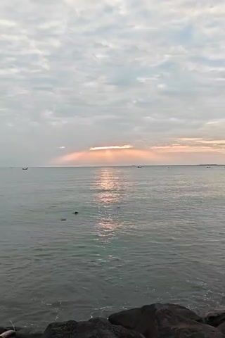 The video shows a sunbeam breaking through the clouds over the ocean. The water is relatively calm, and there are a few boats visible in the distance. A fishing pole enters the frame briefly. The sky is mostly cloudy with a peach hue from the sun. There are large rocks in the foreground at the shoreline.