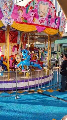 The video pans across an amusement park area. In the foreground, a carousel with "My Little Pony" characters is visible. Several blue and white horses are on the carousel, with a bright blue horse in the center. Behind the carousel, there's a tall, red and blue rocket-shaped ride. Further in the background, other amusement park rides and structures are visible, including what appears to be a Ferris wheel. The ground is covered in a blue and yellow checkered matting. Several people are walking around the park. The sky is clear and blue, suggesting it is daytime. A sign with Chinese characters is posted on the carousel's railing.