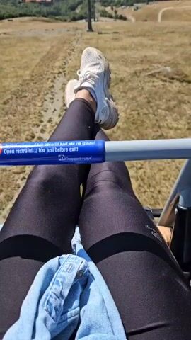 The video begins with a close-up shot of a person's legs, wearing black leggings and white sneakers, resting on a ski lift bar. The bar has a blue stripe with white text, some of which is legible: "restrani... bar just before exit!". The camera then pans up to reveal the wider view from the ski lift. The ski lift is moving uphill, over a dry, grassy mountain landscape. In the distance, several ski lift towers and chairs are visible, as well as winding mountain roads and scattered buildings. The sky is clear and blue, and rolling green hills and mountains dominate the background. The overall impression is of a scenic ride on a ski lift during warmer months, possibly summer or early autumn, given the dry grass.