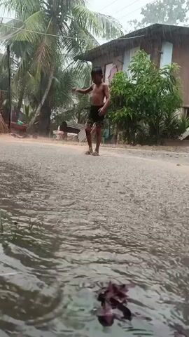 A low-angle shot captures a young boy, shirtless and wearing dark shorts, standing in a large puddle on a rural dirt road during heavy rain. He then runs away from the camera, disappearing from view. The road is visibly wet with numerous puddles and flowing water. In the background, a small, elevated wooden house with a corrugated metal roof is partially visible, surrounded by lush green foliage and palm trees. The continuous falling rain creates ripples on the water's surface.