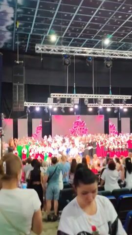 The video shows an indoor event with a large group of people on a stage, likely a performance or a gathering. The stage is lit with bright, pinkish lights and features a backdrop with a large, stylized red design that appears to be a tree or a pyramid shape. There are rows of chairs in the foreground, occupied by an audience. The ceiling is a grid of lights and panels. The overall atmosphere appears to be festive and celebratory. People on stage are dressed in various outfits, including some in red and green, suggesting a coordinated effort. The audience members in the foreground are casually dressed. The scene is brightly lit, suggesting it's daytime or a well-lit indoor venue. The presence of stage lighting, sound equipment (speakers on the side), and a large seated audience indicates a formal event such as a concert, play, or awards ceremony. There is no visible text. The location context of Mykolaiv, Ukraine, provides a geographical setting for the event. The mood of the attendees on stage and in the audience appears to be engaged and positive.