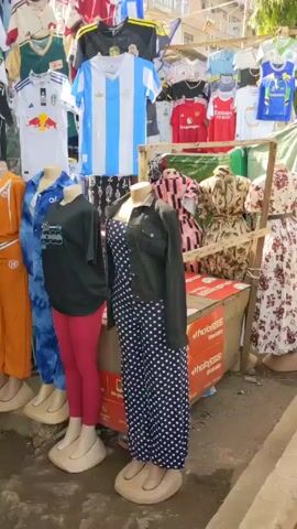 The video depicts a bustling street scene with several vendors displaying their wares along the sidewalk. There's a clothing stall with jerseys and dresses on mannequins, a stall selling phone cases, and another one with jewelry and trinkets.  A man in a white shirt and blue jeans is arranging items at the phone case stall, while a woman in a patterned dress browses the jewelry. A blue motorcycle is parked near the phone case stall. The environment appears to be a sunny outdoor market with a mix of pedestrians and vehicles passing by.
