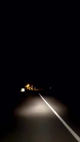 A car drives down a dark road at night, illuminated by its headlights. A white line marks the right side of the road. In the distance are some buildings with yellow lights. The sound of wind can be heard.