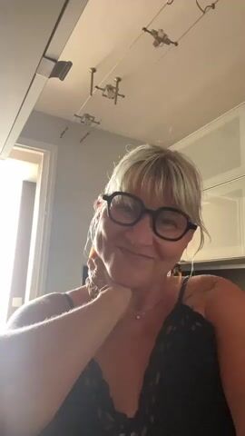 A woman with blonde hair and glasses smiles at the camera, touching her neck. She then adjusts her hair and earrings. She is wearing a black lace top. The background appears to be an indoor setting with a ceiling fixture and a window.