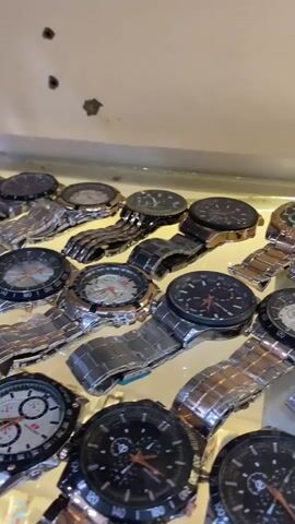The video shows a display of multiple wristwatches. The watches are of various designs, with metal straps in silver and rose gold colors, and watch faces in black and white. Some watches have multiple smaller dials on their faces, indicating chronograph functions. The watches are laid out on a reflective surface, possibly a mirror or glass, under artificial lighting. The background is blurred but appears to be a retail environment, possibly a store or market stall. The camera is positioned above and slightly to the side of the display, panning across the collection of watches. There are no people visible in the video. The audio contains a voice speaking in Indonesian, saying "Ini bisa bawa ke sungai" which translates to "This can be taken to the river."