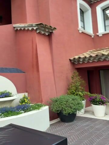 The video begins with a close-up of the exterior of a red building, featuring a decorative tiled roof and a white fountain with blue flowers. The camera then pans out to reveal an outdoor patio area with a large white umbrella providing shade. The patio is furnished with wooden benches, a coffee table, and several round tables covered in white tablecloths. Beyond the patio, there is a well-manicured garden with trees, hedges, and flowers. In the distance, figures can be seen walking across the lawn. The camera continues to pan, showcasing more of the garden and the trees surrounding the patio.