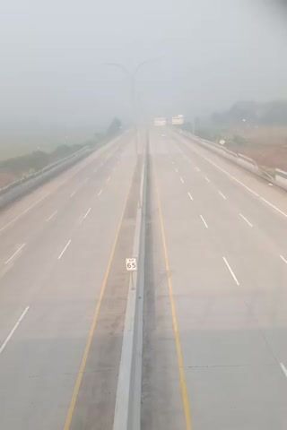 The video shows an aerial view of a highway on a foggy day. The highway is empty except for a few cars in the distance. The lanes are marked with white lines and there is a yellow line dividing the traffic. There is a speed limit sign with the number 65 on it. The fog obscures the horizon and the surrounding landscape.