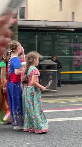 A person's hand holding a phone is in the foreground, capturing a street scene. In the middle ground, a person in a colorful Indian outfit, with a braid and holding a fan, walks away from the camera. Further in the background, a bus stop with a yellow bench is visible. Several other people are walking in the same direction as the person with the fan, some in traditional Indian clothing, and others in more casual attire. The environment appears to be an urban street with a bus stop and buildings in the background. The lighting suggests it is daytime, possibly overcast.
