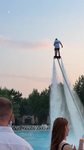 At 00:00, a single person stands on a hydroflight device over a body of water, with two streams of water shooting downwards. A crescent moon is visible in the pale sky. Trees form a backdrop behind the person and the water. In the foreground, the backs of two people's heads are visible, looking towards the hydroflight device. At 00:01, the person on the device begins to be joined by another person. By 00:02, the second person has fully joined the first, and they are now embracing. The two figures continue to hold each other as they are propelled by the water jets. Throughout the video, the setting appears to be an outdoor recreational area with a pool and some covered seating structures visible in the background, with a gentle sunset sky.