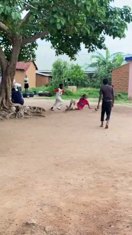 The video shows a group of children playing soccer on a dirt ground in what appears to be a rural or semi-rural setting in Riyom, Nigeria. The scene is outdoors under natural daylight, with an overcast sky visible in the background.

In the foreground, a boy wearing a white shirt and red shorts is seen falling to the ground at the beginning of the clip, then quickly getting back up. Another boy, dressed in a dark brown t-shirt and black pants, stands nearby, observing the play. A third boy in a red t-shirt sits on the dirt ground further back on the left.

After getting up, the boy in the white shirt energetically kicks the soccer ball across the dusty field. Simple, single-story houses, constructed with what appears to be mud bricks and some with painted walls (one pink), are visible in the background. Lush green trees and vegetation dot the landscape, including a large tree providing shade on the left side of the frame. Towards the end of the clip, the boys start to walk away, suggesting a break in their game or the ball has gone out of frame. The overall atmosphere is one of casual, unstructured play in a community setting. No visible text is present in the video.