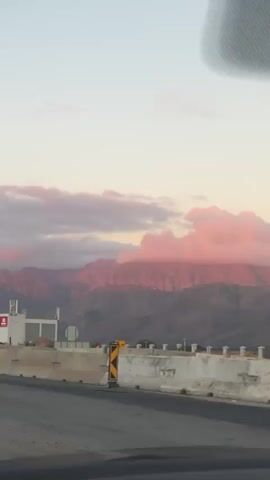 The video shows a car driving down a road lined with yellow and black striped traffic signs. In the background, there is a mountain range with a pink hue, likely due to the sunset or sunrise. The sky is a mix of light blue and pink clouds. On the left side of the road, there's some construction activity with concrete barriers and a building with a white and red sign. The road itself is relatively new and marked with white lines.