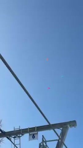 The video shows a view of a ski lift tower and its cable against a clear blue sky. Three distant colorful objects, possibly kites or flags, are visible in the sky. The camera moves slightly, tracking the position of the objects in the sky in relation to the ski lift infrastructure. The number "6" is clearly visible on the ski lift tower structure.