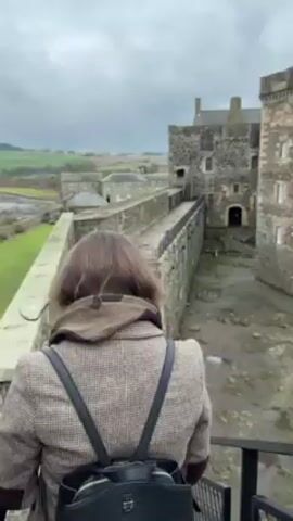 A woman is standing on a set of metal stairs, looking down at a medieval castle. She turns her head to the side and looks towards the camera. She is wearing a brown tweed coat, a brown hoodie, and a black backpack. The castle is made of grey stone and has a walkway along the top of the walls. There is a field in the background and the sky is cloudy.