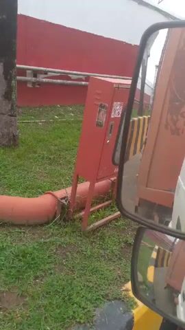 The video shows an outdoor scene with a red wall and a white structure in the background. In the foreground, there is a red fire hose cabinet on metal legs, with a large orange pipe extending from the ground towards the cabinet. A palm tree is visible to the left of the scene. A side mirror of a vehicle reflects part of the scene, including a yellow and black striped barrier and the side of an orange vehicle. The area is covered with grass. Barbed wire is visible along the top of the red and white wall.