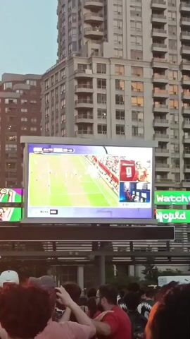 A group of people are gathered outdoors, facing a large digital screen displaying a soccer match. The screen is situated on a structure above the crowd. To the left of the main screen, another smaller screen shows people cheering, with images of soccer balls and faces. In the background, a tall modern building rises into the sky. The sky appears to be overcast or at dusk. The crowd is diverse, with individuals of various ages and ethnicities. Some people are holding up their phones, presumably to record or take pictures of the event. The overall atmosphere seems to be one of excitement and shared anticipation.