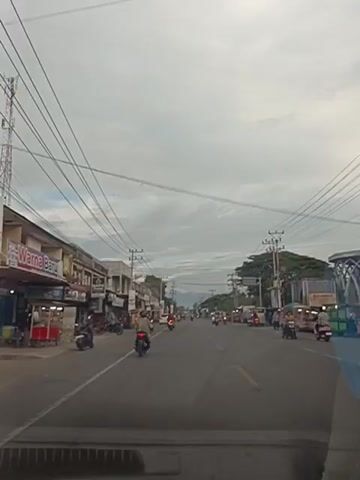 A street scene captured from a moving vehicle. On the left, a row of buildings with shops line the road. Signs on the buildings include "Karna Baru" and "MAFIZ FASHION". Several motorcycles are parked in front of the shops. A red food cart is also visible. Traffic on the road consists of motorcycles and a few cars. On the right side of the road, more shops and stalls are present, with some trees and vegetation in the background. Utility poles and power lines crisscross the sky. The sky is overcast with clouds. The perspective suggests the vehicle is moving forward along the road.