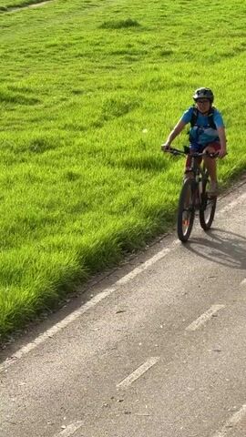 This media file features a person riding a mountain bike on a paved path next to a grassy field. The cyclist is wearing a blue t-shirt, red shorts, a helmet, and glasses, and is carrying a blue backpack. The sun is shining, casting long shadows from the cyclist and their bike, suggesting it is late afternoon or early morning. The lush green grass of the field contrasts with the grey asphalt of the path, creating a vibrant natural scene. The path appears to be a dedicated bike lane or trail. The cyclist is looking towards the camera and appears to be smiling, indicating a pleasant and enjoyable ride. The backdrop of the grassy field suggests an outdoor recreational area, possibly a park or a scenic route. The lighting and the overall atmosphere convey a sense of activity and outdoor enjoyment. There is no visible text in the media file.