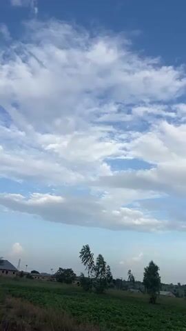The video shows a mostly clear blue sky with scattered white clouds. The clouds appear to be moving gently. Below the sky, there is a landscape with green fields, trees, and some buildings. The overall impression is of a peaceful rural setting on a day with fair weather.