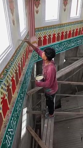 A man is painting the interior of a building. He's standing on wooden planks that are resting on scaffolding. He is wearing a maroon shirt, black pants, and sandals. He has a small bag slung over his shoulder and is holding a container in his left hand. He is using a brush in his right hand to paint a green band that has white Arabic calligraphy on it. The wall below the green band has alternating red and yellow decorations. Above the green band is a red and green pattern. The room has multiple windows. The interior is still under construction. The man is focused on his task.