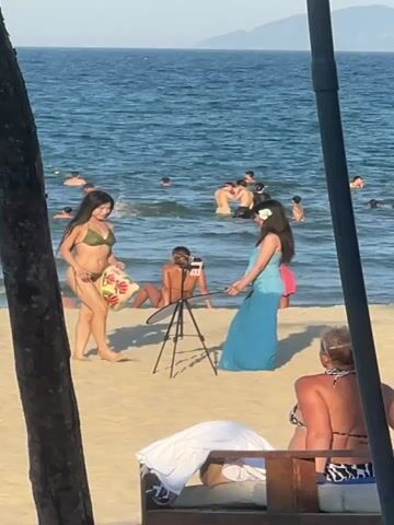 This media file captures a beach scene in Đà Nẵng, Vietnam, during daylight hours under clear skies. The primary focus is on a group of people on the sand near the ocean.

In the foreground, partially obscured by foliage, a woman with blonde hair sits on a wooden lounge chair, wearing a bikini. Behind her, a sandy beach stretches towards the water, where several people are swimming and enjoying the ocean.

The middle ground features two young women on the beach. One, in a green bikini, holds a conical hat and appears to be posing. The other, in a long, flowing blue dress, holds a reflector. A tripod with a camera is set up between them, suggesting a photoshoot is in progress. Another woman is visible sitting on the sand behind them, facing away from the camera.

The ocean is a vibrant blue, with gentle waves. The horizon is visible, with a hazy mountainous landscape in the distance. The overall atmosphere is relaxed and summery, indicative of a typical day at a popular beach destination. No text is visible in the media file.