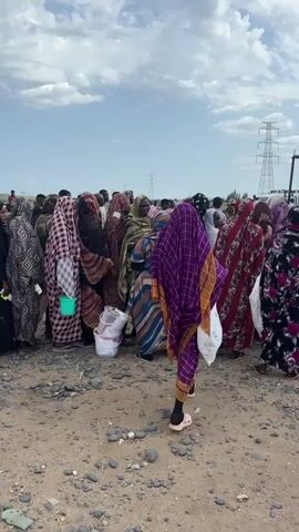 This media file depicts a group of people, predominantly women, gathered outdoors in what appears to be a semi-arid environment. They are dressed in colorful, traditional clothing, including hijabs and abayas, suggesting a cultural context. Some individuals are carrying bags or buckets, hinting at the collection or distribution of goods.

The scene is set on a dirt and gravel ground, with scattered stones. In the background, several large white sacks are visible, stacked beneath a large, crimson tarp supported by poles. This suggests an organized distribution point for supplies, possibly food or water. The presence of high-voltage power lines and pylons in the distance indicates the proximity of infrastructure, but the overall setting seems somewhat remote, with sparse vegetation.

The sky is partly cloudy, with a bright, diffused light, suggesting it is daytime, likely late morning or early afternoon. There are no clear indicators of specific emotions, but the gathering of people suggests a communal activity or event, possibly related to aid distribution or a marketplace. The overall atmosphere is neutral, focused on the practicalities of the scene.

The image does not contain any visible text. The location context provided is Ad-Damazin, Sudan. This suggests the scene is likely related to humanitarian efforts or community activities within that region.