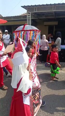 This video shows a parade or procession taking place outdoors on a sunny day. Several children and adults are participating, many wearing red and white outfits. Some individuals are carrying umbrellas, one large umbrella is decorated with colorful ribbons. In the foreground, a child in a red and white costume is walking away from the camera. To the left, a girl in a red and white uniform is walking forward, holding a piece of cardboard. Further back, several adults in red and white attire are also walking. In the background, there are buildings and what appears to be a fence or barricade. The overall atmosphere suggests a festive community event.