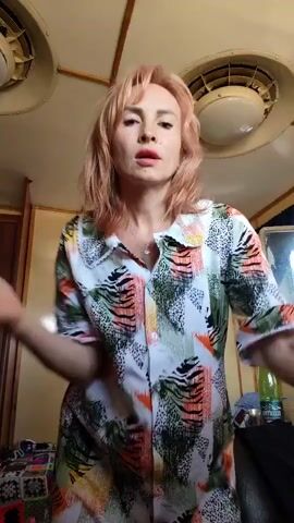 A woman with short, peachy-colored hair stands in what appears to be a cabin or a small room. She is wearing a colorful, button-down shirt with a bold, abstract pattern featuring shades of green, orange, white, and black. Her arms are initially crossed in front of her, and her mouth is slightly open. She then moves her hands, touching her hair and then bringing them down to her sides. She leans forward, bringing her face closer to the camera with a subtle smile. The room has a wooden paneled ceiling with two circular light fixtures. In the background, a dark wooden door is visible on the left. To the right, a green bottle and some other objects are on a surface, and a partial view of a mirror can be seen. The overall lighting suggests it might be daytime, but the indoor setting makes it difficult to determine the exact time or weather conditions. There is no visible text in the media.
