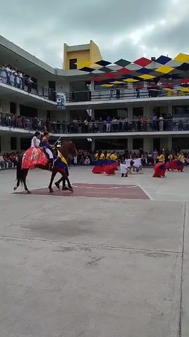 This video captures an outdoor cultural performance taking place in a courtyard in Cabudare, Venezuela. In the foreground, a person rides a brown horse, adorned with a vibrant, multi-colored skirt featuring red, white, blue, and yellow patterns. Several other individuals are on the ground, some dressed in traditional attire with long skirts in the colors of the Venezuelan flag (red, yellow, and blue). One person is holding a large Venezuelan flag. A few other people are seen crouching or kneeling in the central performance area.

The performance is watched by a significant audience, with spectators lining the ground level and gathered on the balconies of the surrounding two-story buildings. These buildings feature a modern architectural style. Above the courtyard, a decorative canopy of square panels in red, yellow, blue, black, and white provides shade and adds to the festive atmosphere. The sky is overcast, indicating a cloudy daytime. The scene suggests a lively community or school event celebrating Venezuelan culture. No explicit text is visible in the video.