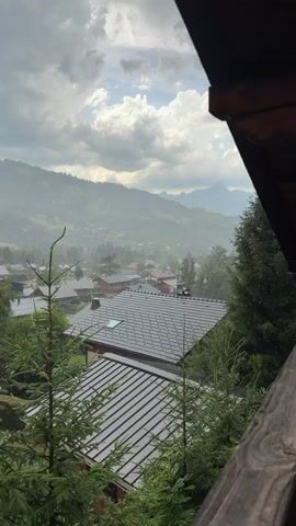 This is a vertical video clip showing a picturesque view from a balcony overlooking a village nestled in a valley. The scene is dominated by a heavy rainfall, with visible streaks of rain falling through the air and glistening on the dark, sloped rooftops of the houses below. 

In the foreground, several tall fir trees with lush green needles are positioned on either side of the frame, adding depth and a natural element to the composition. The balconies of what appears to be a wooden chalet are visible on the right side of the image, suggesting the viewpoint is from an elevated position within a mountain resort.

Beyond the immediate cluster of houses, the valley unfolds with more residential buildings scattered amidst dense greenery. Rolling hills and mountains form the backdrop, their outlines softened by the rain and mist, creating a slightly hazy and atmospheric effect. The sky is dramatic, with a mix of dark, heavy rain clouds and patches of bright, diffused sunlight breaking through, hinting at a dynamic weather system. The overall mood is tranquil and somewhat melancholic, evoking the feeling of a cozy, rainy day in a mountain town. The time of day is likely afternoon or early evening, given the diffused light. No visible text is present in the video.