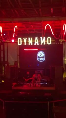 The video shows a DJ setup in a dimly lit club. Red neon lights are visible above the stage, spelling out the word "DYNAMO" in large letters. A screen behind the DJ booth displays the "DYNAMO" logo, which features a stylized animal. Two DJs are present behind the turntables, one in the foreground and another behind them. The overall ambiance suggests a live music or clubbing event.