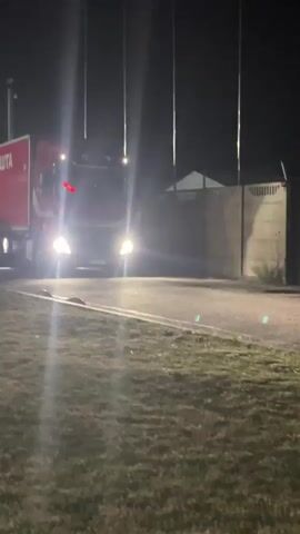 A red semi-truck with white lettering on its trailer drives on a paved road at night. The truck's headlights are on, illuminating the road and a grassy area in the foreground. A concrete wall with a fence on top is visible behind the truck. The truck appears to be turning.