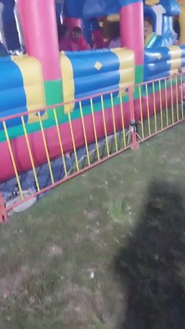 The video shows a large, colorful inflatable bounce house at night. It is primarily blue, pink, and yellow, with cartoon characters on top, including a white rabbit and a yellow character with a flower-like head. A metal barrier fence with yellow and red poles is in front of the bounce house, separating it from a grassy area. Inside the bounce house, children can be seen playing. One child in a pink shirt is visible near the right side of the bounce house. The lighting is dim, suggesting it is evening or night.