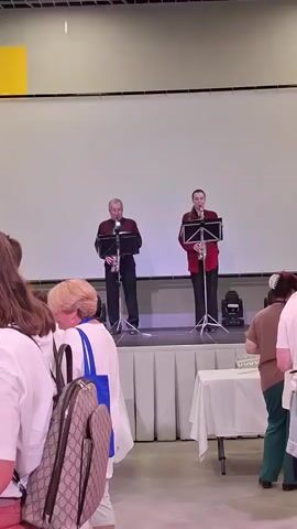 Two men are playing saxophones on a raised stage in front of a large white screen. They are both wearing red shirts and black pants. The man on the left is older and has gray hair. The man on the right is younger and has brown hair. There are several people in the foreground, some of whom are looking at the musicians. There are also tables with white tablecloths in front of the stage. The environment appears to be an indoor venue, possibly a conference hall or auditorium, with a light-colored floor. Stage lighting is visible on the left and right sides of the stage.