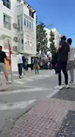 The video captures a bustling street scene in Vejer de la Frontera, Spain, during daylight hours. The setting features a paved street bordered by multi-story white buildings with balconies, typical of Southern Spanish architecture. The sky is clear and blue, indicating sunny weather.

Numerous people are present, both in the foreground and further down the street. They are dressed in casual attire, such as t-shirts, shorts, and trousers. On the far left, an individual holds a vertical object, possibly a flag or banner, which appears to have red and yellow colors. Other people are seen walking across the frame or standing in groups, suggesting a social gathering.

A significant number of people are concentrated further along the street, implying a public event, festival, or celebration is underway. The overall atmosphere appears lively and engaged. Air conditioning units are visible on the facades of the buildings. No discernible text is visible within the frame.
