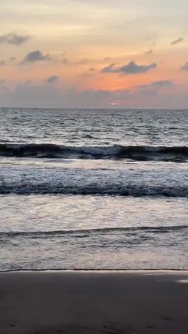 The video shows a sunset over a beach. The sky is filled with clouds tinged with orange and pink. The ocean has gentle waves breaking on the shore. The sand in the foreground is dark. There are no people or objects visible in the frame. The video is very short, lasting only a few seconds.