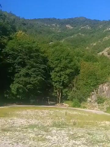 The video shows a landscape scene with a road in the foreground and a rocky, tree-covered mountain in the background. The sky is clear and blue. The road is made of dirt and gravel. The mountain is steep and covered in dense green trees and grey rocks. The sun is shining, casting shadows on the mountain. The video pans slowly across the scene, giving a panoramic view of the landscape. There are no people or vehicles visible in the video. The overall impression is one of natural beauty and tranquility.