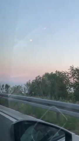 The video shows a view from inside a car, looking out the passenger window and into the passenger side mirror. The sky is a pale blue with streaks of pink and purple, indicating either sunrise or sunset. There is a guardrail visible along the side of the road, separating the car from a steep drop off overlooking the sea. The sea is a hazy blue and stretches to the horizon. Small trees and shrubs grow on the rocky terrain along the roadside. A speed limit sign showing "60" is visible in the distance on the left side of the road. The interior of the car is reflected in the side mirror, including what appears to be the headrest of the driver's seat and a person's hand resting on the steering wheel. The video appears to be shot while the car is in motion.