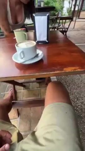The video shows a first-person perspective from someone seated at a wooden outdoor table at a cafe. On the table is a white cup of coffee with latte art, resting on a matching saucer with a small spoon. Next to it is a green mug and a black napkin holder with white napkins. A woman with long brown hair, wearing a white tank top, looks toward the table and is then joined by a waitress who places a second cup of coffee on the table for the woman. The environment is a paved patio with green foliage visible in the background. Other tables and chairs can be seen scattered around the patio. The person filming is wearing khaki shorts.