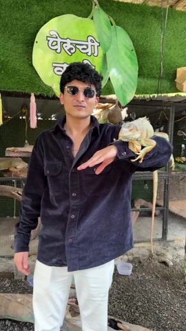 A person with dark, curly hair and sunglasses stands in front of a green backdrop and a caged area. The person wears a dark corduroy button-down shirt and white pants. An iguana is perched on the person's arm. The iguana is light green and orange, with a long tail. The backdrop has a green apple shape with white text in a script font. Sunglasses and a birdhouse are visible in the cage area. The ground is covered in small, dark gravel.