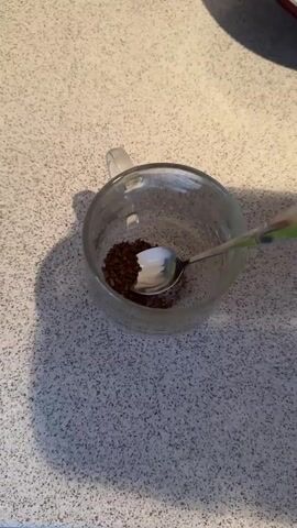 The video shows a clear glass mug on a speckled countertop. Inside the mug, there are dark brown granules, presumably instant coffee, and a silver spoon. Water is being poured from an unseen source into the mug, mixing with the coffee granules. The water is clear and the coffee turns a dark brown as it dissolves. The mug casts a shadow on the countertop to its left.