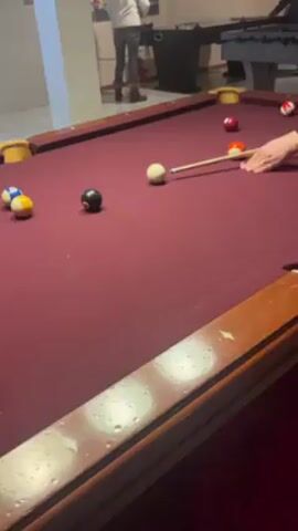 The video shows a person playing pool. The pool table is red and there are several balls on the table, including the eight ball. A person wearing a white sweater and jeans is standing in the background near a vending machine. The camera pans around the table as the player lines up a shot. The player strikes the cue ball.