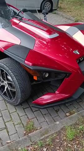 The video shows a red Polaris Slingshot parked on a brick-paved surface. The vehicle has two seats, a windshield, and a single rear wheel. A silver car is parked behind the Slingshot. The scene appears to be outdoors, possibly in a parking lot or driveway, with some greenery visible in the background. The camera pans slightly to show the side of the Slingshot.
