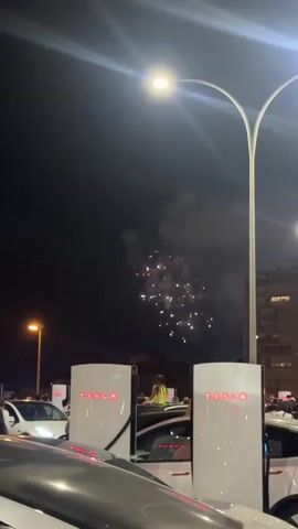 The video opens with a nighttime scene in what appears to be a Tesla charging station. The foreground is dominated by the front of a white Tesla vehicle. In the middle ground, several white Tesla charging stations with illuminated red "TESLA" logos are visible. One charging station has a reflection showing a woman in a yellow top sitting on the roof of a car. Another Tesla vehicle is parked nearby, and to the left, there are more cars and charging stations. The background features a dark night sky with scattered fireworks creating bursts of light and smoke. A street lamp with a bright light shines down from a curved pole in the upper right portion of the frame. The overall environment suggests an outdoor urban setting at night, possibly during a celebration.