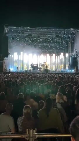 A wide shot captures a concert crowd facing a brightly lit stage. The stage features a band performing with instruments and stage lights. The audience is densely packed, with many people visible from behind, looking towards the stage. The overall atmosphere appears to be a live outdoor event at night.
