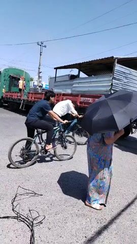 Two individuals are riding on a single bicycle in an outdoor urban environment. The person in the back is wearing a dark blue t-shirt and black pants. The person in the front is wearing a white shirt. They are riding on a paved road. A person holding a black umbrella is standing to the right of the frame. A large truck is parked on the right side of the road. In the background, a train is visible, with several carriages. A white car is parked on the left side of the road. The sky is clear and blue, indicating a sunny day.