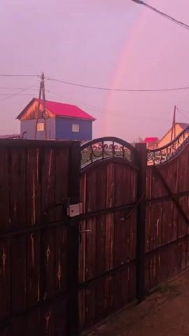 The video displays a shot of a fence with a rainbow in the sky. The fence is made of dark wood with ornate metal details on top. Behind the fence, there are some houses with red roofs. The sky is a soft pink color, suggesting it is either sunrise or sunset. There are also power lines visible in the upper part of the frame. The scene appears to be in a rural or suburban setting.